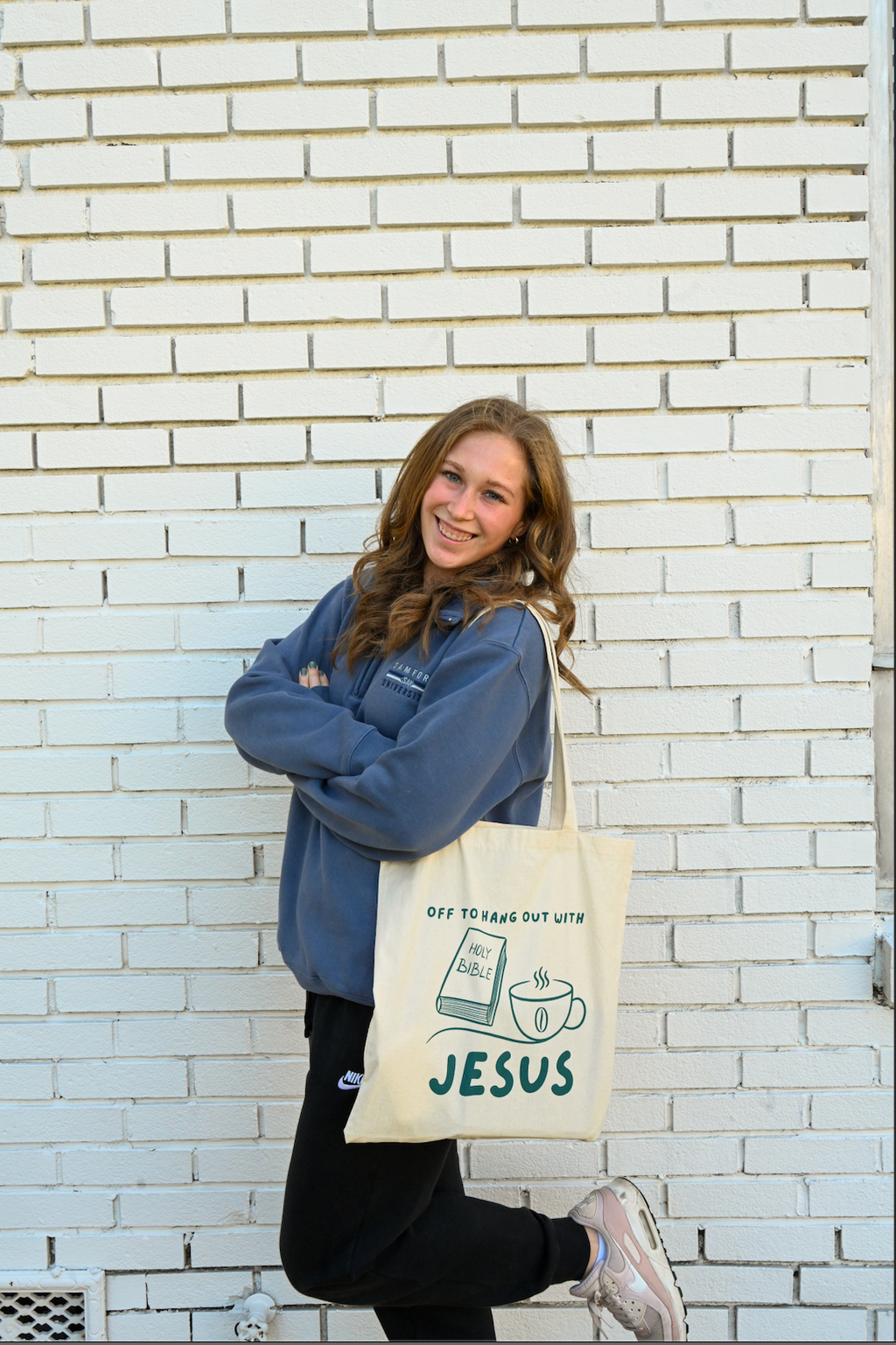Person holding a tote bag with 'Off to hang Out with Jesus' text against a white brick wall.