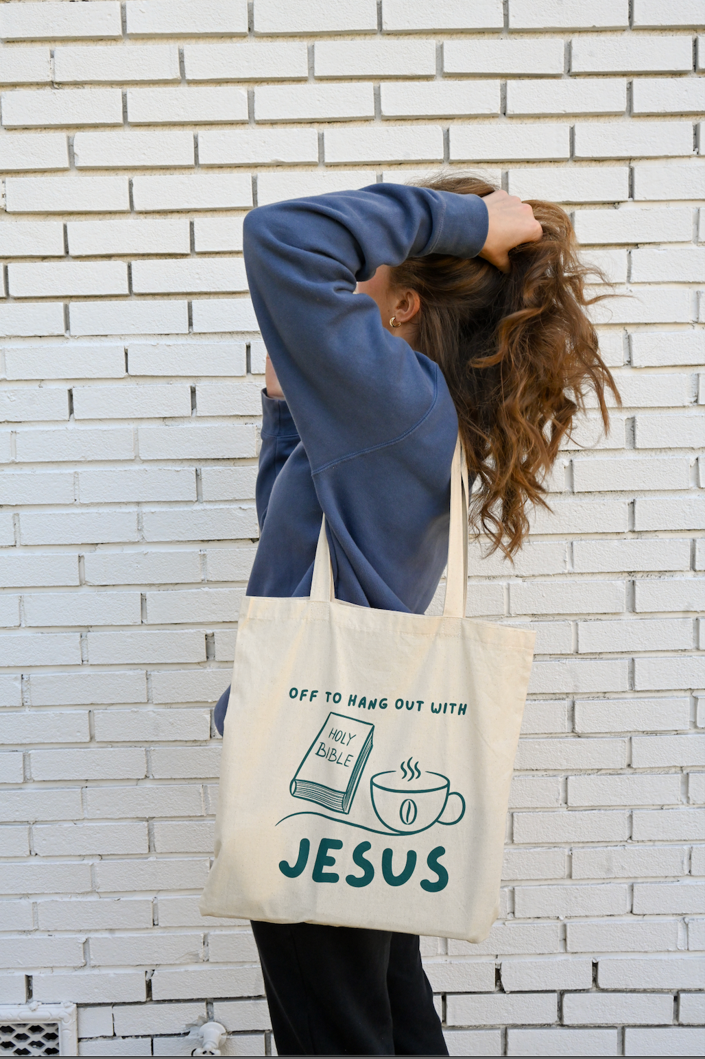 Person holding a tote bag with a christian design against a white brick wall.