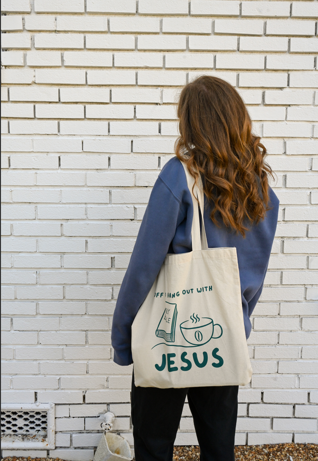 Person holding a tote bag with 'off to hang Out with Jesus' text against a white brick wall.