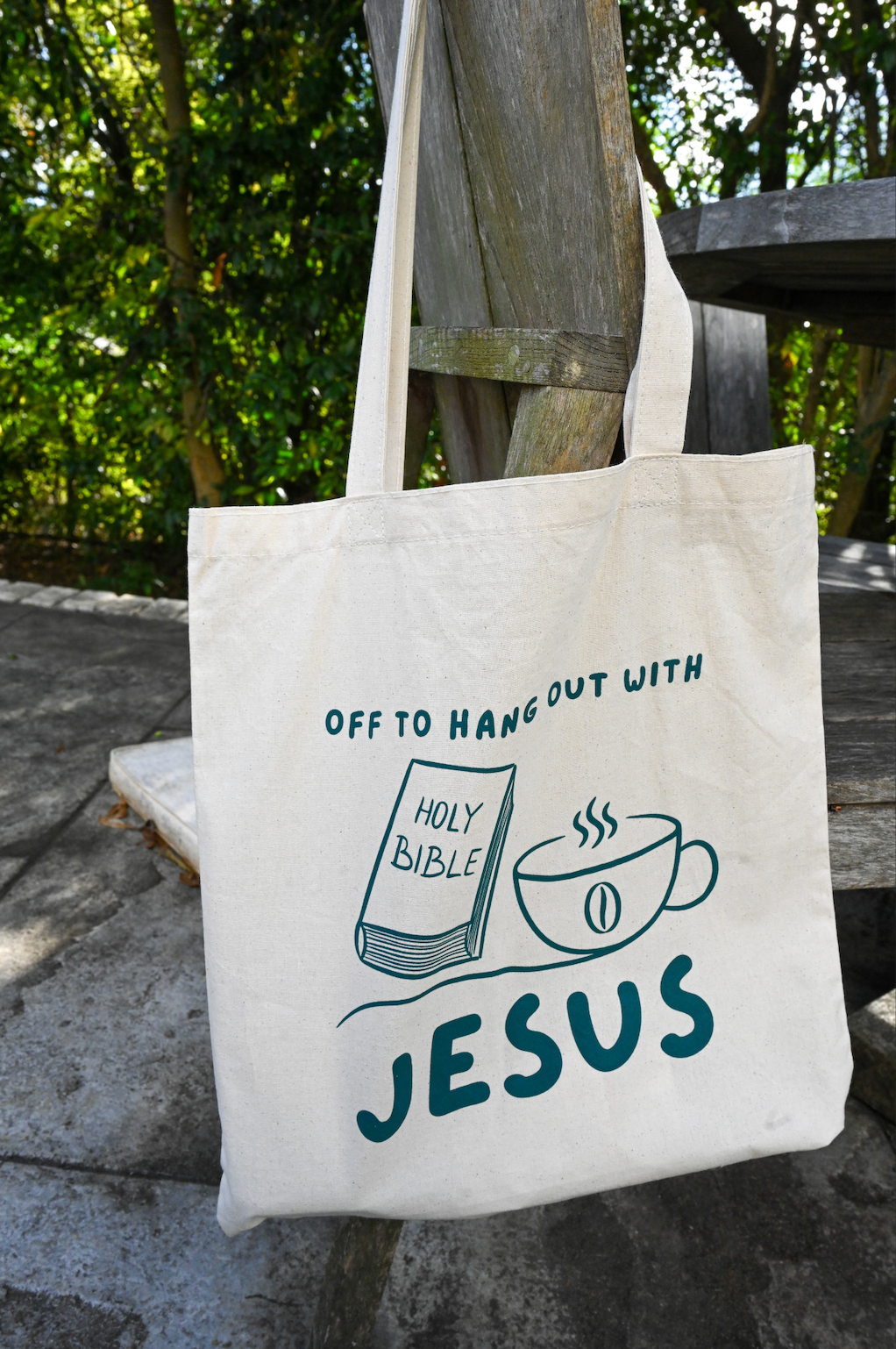 Tote bag with 'Off to hang out with Jesus' design featuring a Bible and coffee cup on a wooden bench.