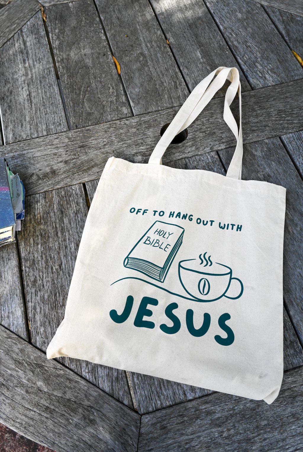 Tote bag with 'Off to hang out with Jesus' text and graphics on a wooden surface